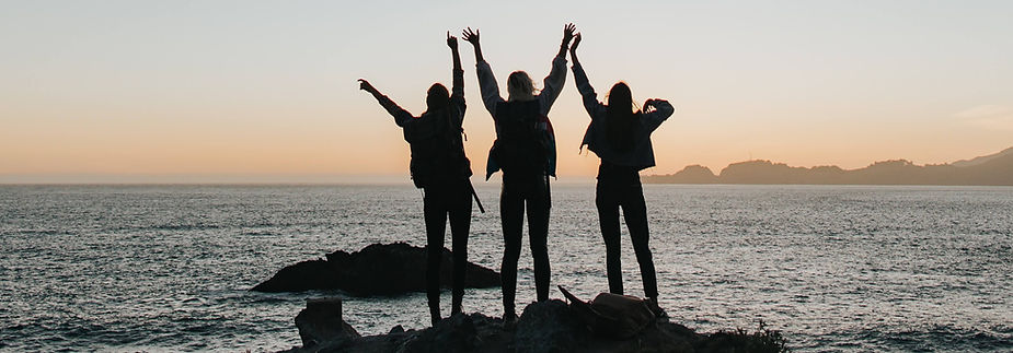 three people with their arms up standing on a rock at the ocean at dusk